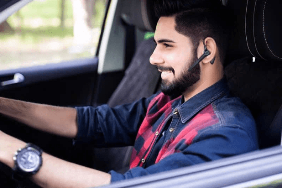 A smiling bearded man wearing a red and black plaid shirt and a wireless Bluetooth earpiece while driving a car.
