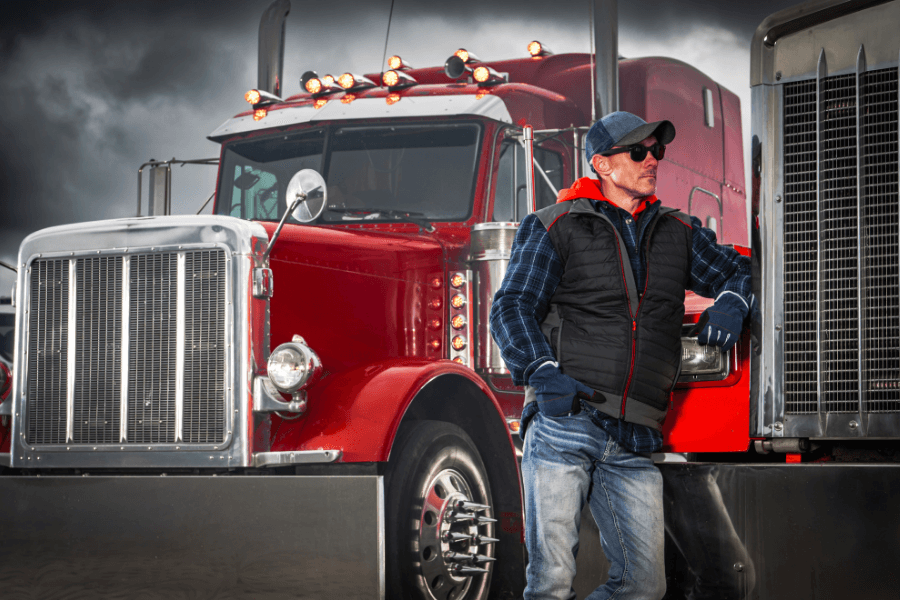 A male truck driver wearing sunglasses, a cap, a plaid shirt, and a vest, leaning against a classic red semi-truck under a cloudy sky.