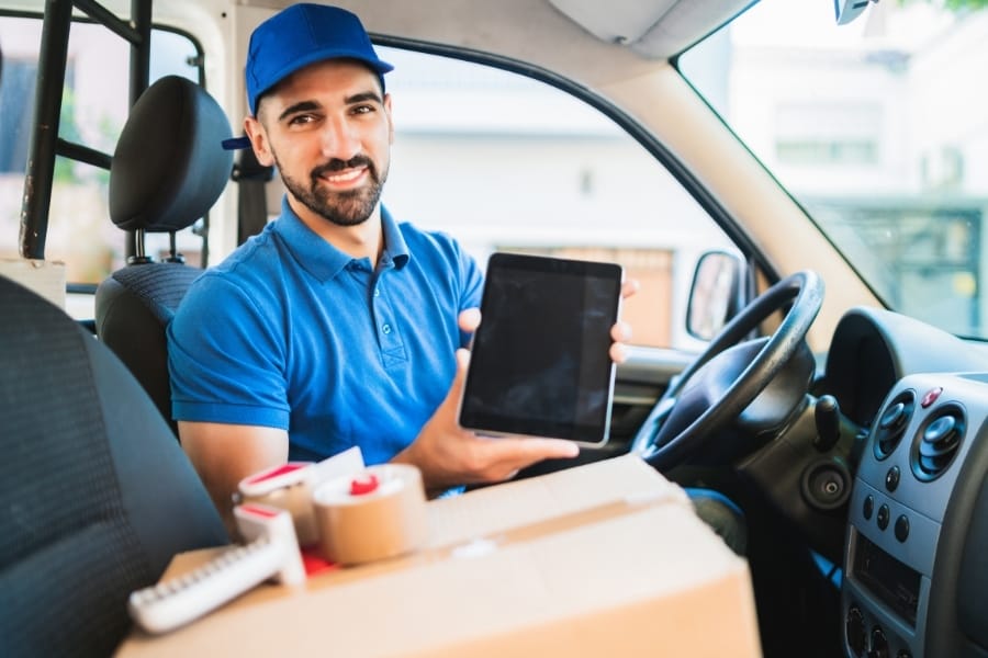 A delivery driver in a blue uniform and cap holding a tablet while sitting inside a delivery van with packages.