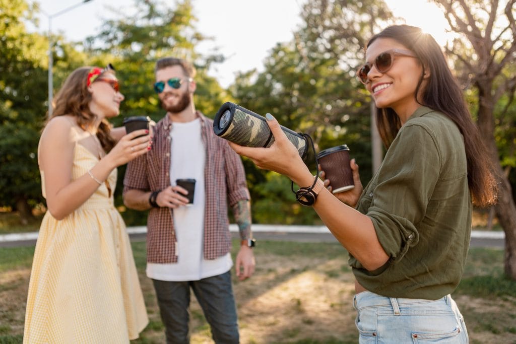 Friends chatting outdoors while carrying a portable Bluetooth speaker and coffee