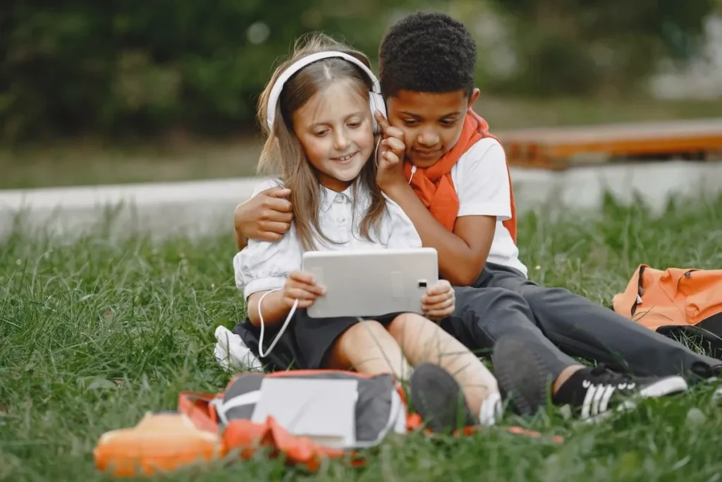 Two children sitting on grass and looking at a tablet, with one wearing headphones.