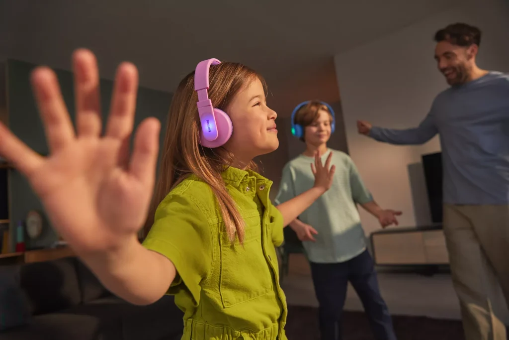 Girl wearing pink Philips TAK4206 headphones while dancing with a boy wearing the blue model and an adult at home.